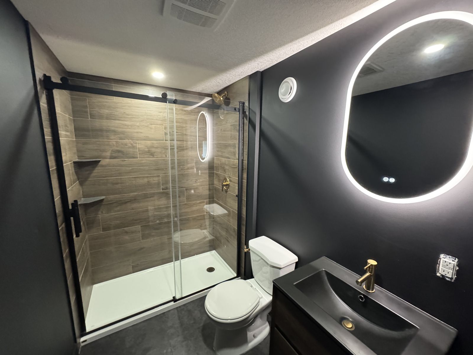 Dark bathroom with black walls, wood-look tile shower, black framed glass and an oval backlit mirror
